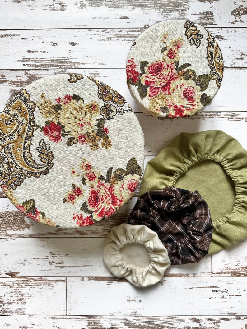 May include: A set of five round fabric bowl covers in various sizes. Two covers feature a cream background with red roses and brown paisley patterns. One cover is solid sage green, one is brown and black plaid, and one is solid cream.