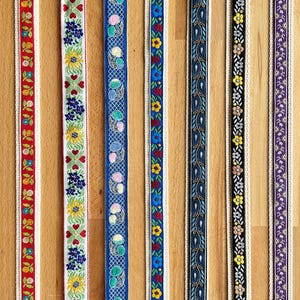 May include: Seven vertical strips of decorative woven ribbon featuring distinct patterns: red floral, red hearts, blue cherries with a grid, blue floral, blue paisley, black floral, and purple floral. Each ribbon has a cream-colored border.