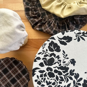 May include: A collection of four round fabric bowl covers with elasticated edges. One cover features a black floral pattern on a white background, two have a brown and black plaid design, and one is a solid light yellow fabric with a gathered texture.