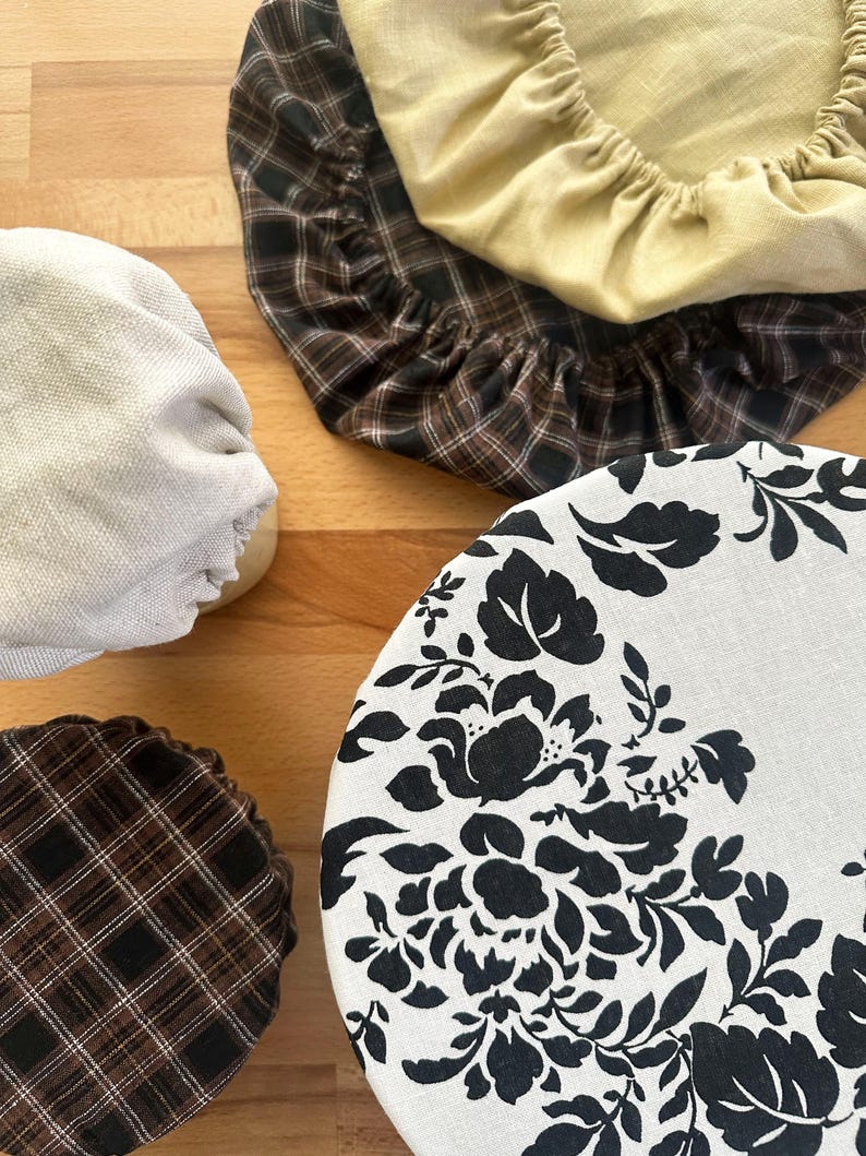 May include: A collection of four round fabric bowl covers with elasticated edges. One cover features a black floral pattern on a white background, two have a brown and black plaid design, and one is a solid light yellow fabric with a gathered texture.