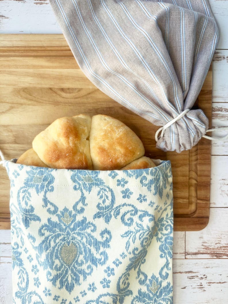 Puede incluir: Un pan dorado se encuentra dentro de una bolsa de pan con estampado azul y blanco. Una bolsa de pan de lino a rayas est&aacute; atada con una cuerda blanca. El pan y las bolsas est&aacute;n sobre una tabla de cortar de madera.