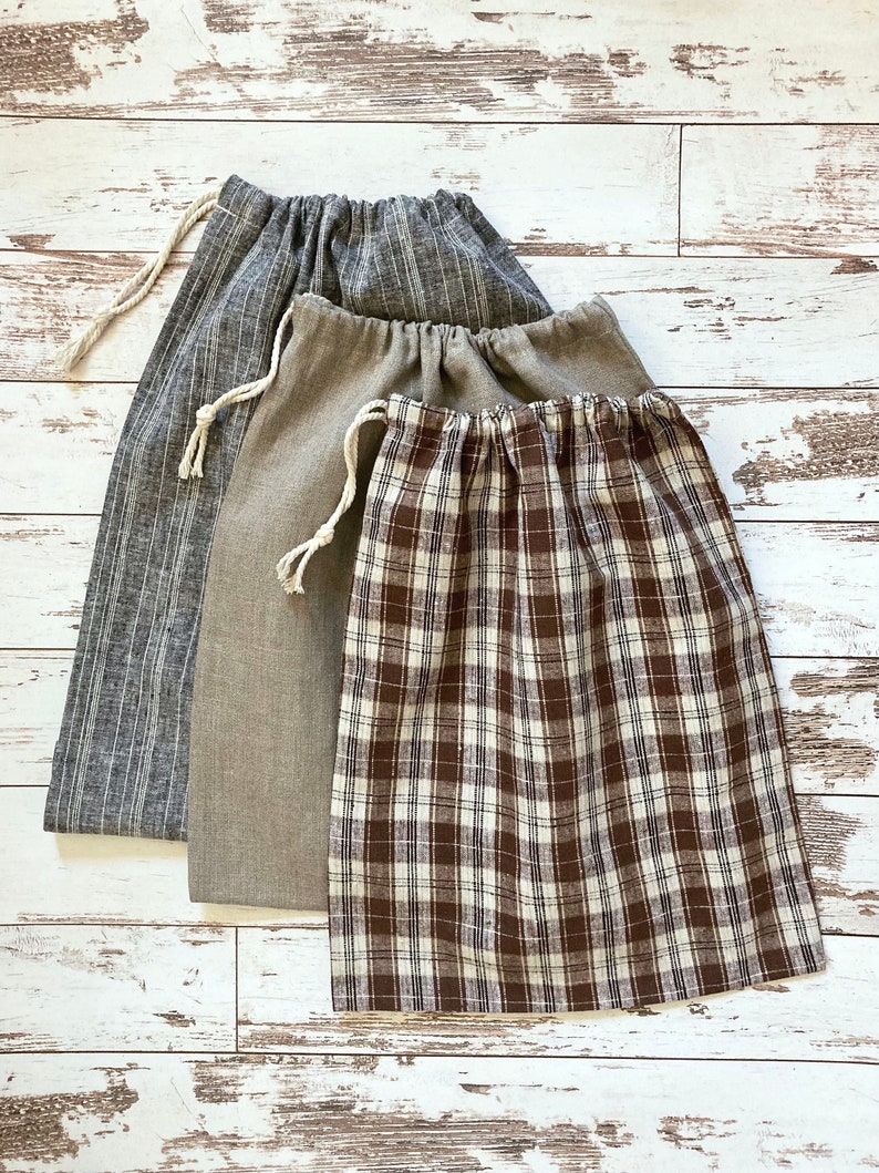 May include: Three drawstring bags in varying sizes and patterns. The largest bag is gray with vertical stripes, the middle bag is solid tan, and the smallest is brown and white plaid. All bags have a natural rope drawstring.