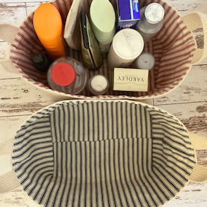 Op de afbeelding: Twee gestreepte opbergmanden van stof. De bovenste mand is gevuld met verschillende toiletartikelen, waaronder een rood-wit gestreepte tube, een groene fles, een blauwe doos, een witte fles met een rood deksel en een witte zeep. De onderste mand is leeg.