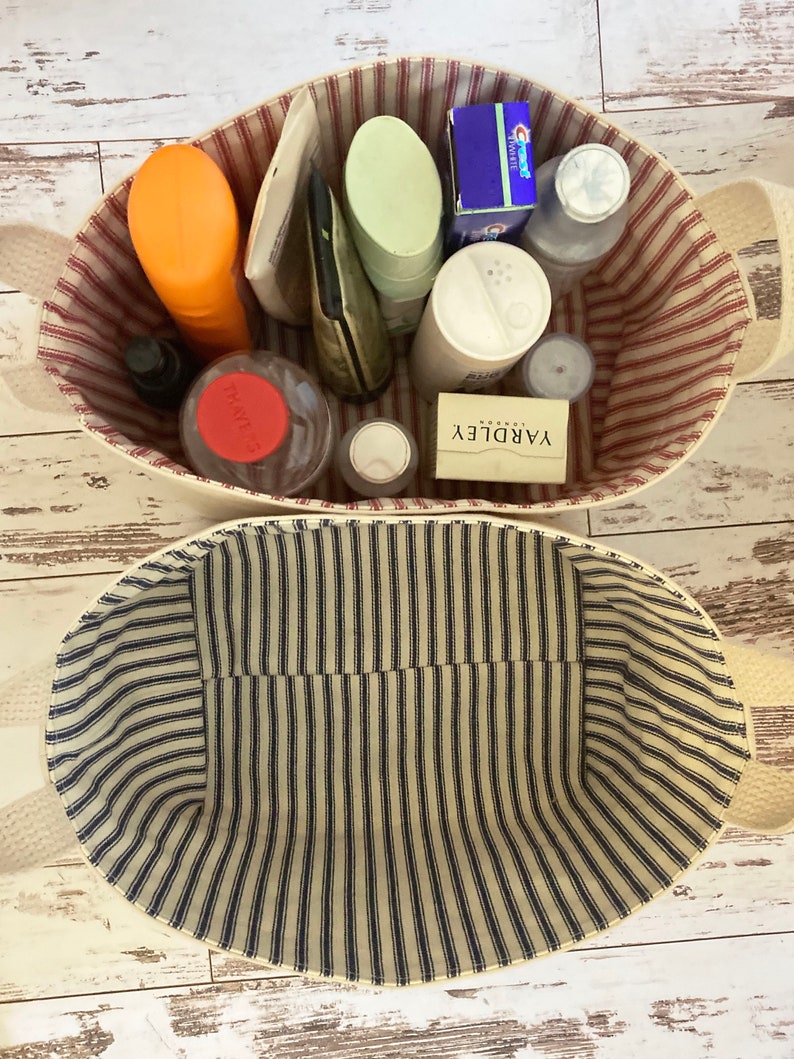 Op de afbeelding: Twee gestreepte opbergmanden van stof. De bovenste mand is gevuld met verschillende toiletartikelen, waaronder een rood-wit gestreepte tube, een groene fles, een blauwe doos, een witte fles met een rood deksel en een witte zeep. De onderste mand is leeg.