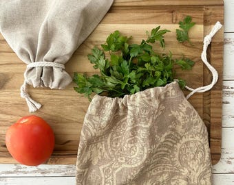 May include: Two drawstring produce bags made of fabric, one plain beige and one featuring a tan and cream damask pattern, are filled with fresh green parsley. A single red tomato sits beside the patterned bag on a wooden cutting board.