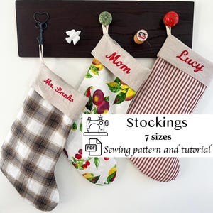 May include: Three fabric stockings hanging from a dark wooden board. The left stocking has a brown plaid pattern, the middle features a fruit print, and the right has red and white stripes. Each has a beige cuff with embroidered names.