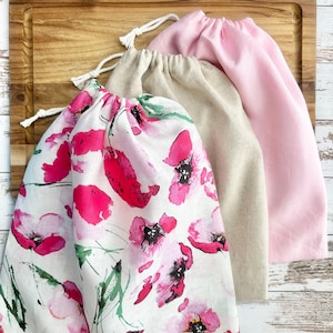 May include: Three drawstring bags featuring a white bag with a pink and green floral watercolor print, a solid beige bag, and a solid light pink bag. Each bag is made of fabric and includes a white cord drawstring closure at the top opening.