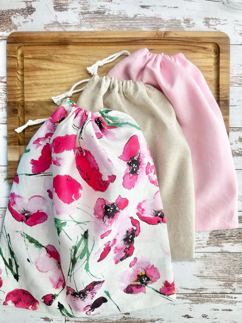 May include: Three drawstring bags featuring a white bag with a pink and green floral watercolor print, a solid beige bag, and a solid light pink bag. Each bag is made of fabric and includes a white cord drawstring closure at the top opening.