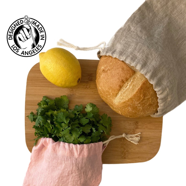 May include: A wooden cutting board with a loaf of bread in a beige drawstring bag, a yellow lemon, and a bunch of cilantro in a pink drawstring bag. The image includes a circular logo that says "Designed Made in Los Angeles."