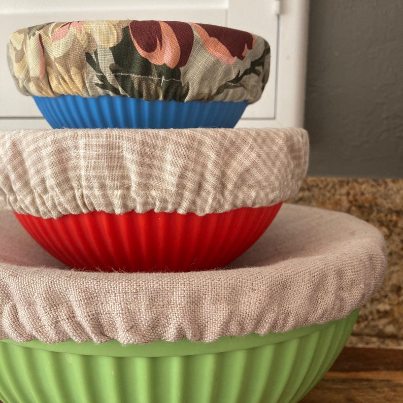 May include: A stack of three ribbed bowls in blue, red, and light green, each topped with a fabric cover. The top bowl has a floral print, the middle bowl has a beige checkered pattern, and the bottom bowl has a solid beige fabric cover.