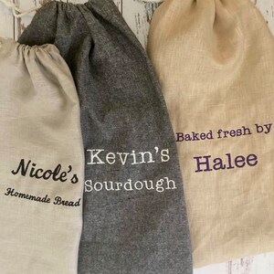 May include: Three linen bread bags with drawstring closures. One bag is gray with "Kevin's Sourdough" embroidered in white. Another is beige with "Baked fresh by Halee" in purple. The third is beige with "Nicole's Homemade Bread" in black.
