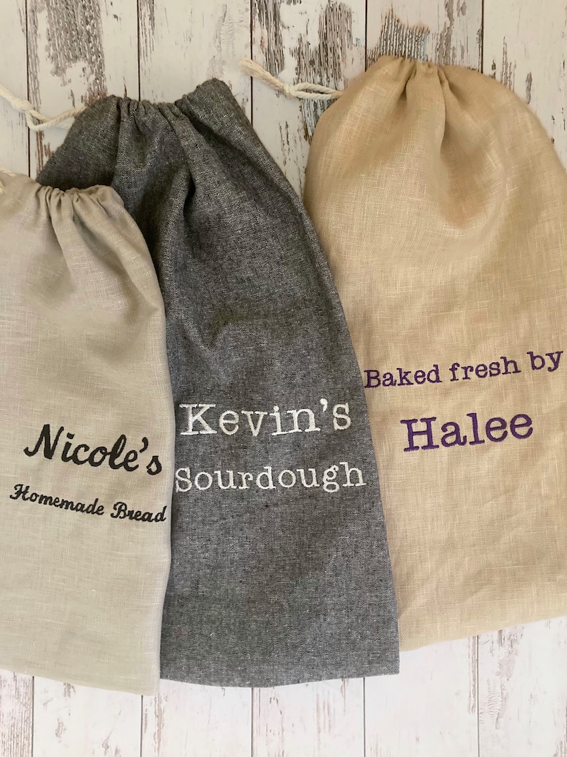 May include: Three linen bread bags with drawstring closures. One bag is gray with "Kevin's Sourdough" embroidered in white. Another is beige with "Baked fresh by Halee" in purple. The third is beige with "Nicole's Homemade Bread" in black.