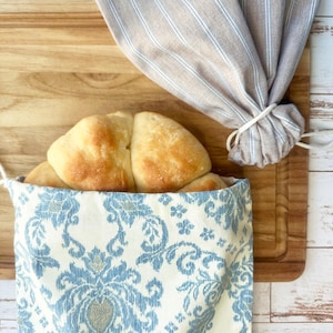 May include: A loaf of golden-brown bread rests inside a blue and white patterned bread bag. A striped linen bread bag is tied with a white string. The bread and bags are on a wooden cutting board.