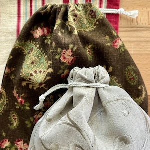 May include: A dark brown drawstring bag with a floral and paisley pattern, a light gray drawstring bag with embroidered swirls, and a rectangular fabric piece featuring red, blue, and cream vertical stripes with a white drawstring closure.