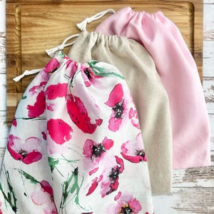 May include: Three drawstring bags on a wooden cutting board. The front bag is white with a pink floral pattern. The middle bag is beige, and the back bag is light pink. All bags have white drawstrings.