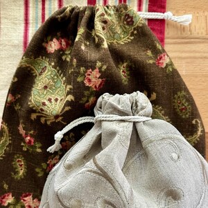 May include: Three drawstring bags with various fabric patterns. The largest bag is brown with a floral and paisley design. The middle bag is a light gray with an embroidered pattern. The background is a striped fabric.