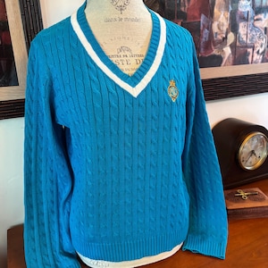 May include: A turquoise cable-knit sweater with a V-neck and white trim. The sweater features a small emblem on the chest and is displayed on a mannequin. The sweater is a classic style.