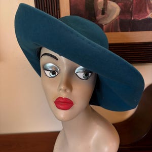May include: Teal-colored wide-brimmed hat on a mannequin head with painted features. The hat has a rounded crown and a wide, downward-sloping brim. The mannequin has red lips and silver eyeshadow. The hat is the focal point, suggesting a vintage or fashion theme.