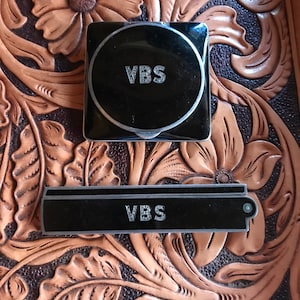 Puede incluir: Dos objetos rectangulares negros con las letras "VBS" en letras plateadas. Los objetos están sobre un fondo de cuero marrón con un diseño floral.