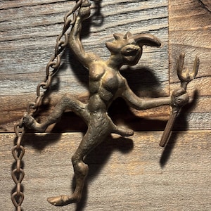 May include: A bronze-colored metal figurine of a horned, humanoid creature with a tail and a pitchfork. The figure is suspended from a chain and is posed in a dynamic, almost dancing position. The background is a wooden surface.