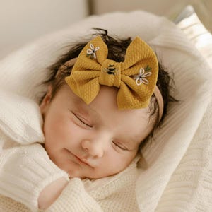 May include: A sleeping baby wearing a mustard yellow bow headband. The bow features embroidered bee and floral details. The baby is wrapped in a white blanket.