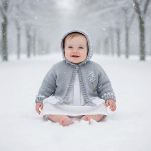 May include: A baby wearing a gray hooded cardigan with a white floral design and a white dress sits in the snow. The cardigan has white trim and is buttoned in the front. Snowflakes fall around the baby.