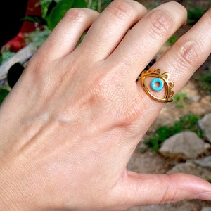 May include: Gold ring with a blue evil eye design. The ring is worn on a person's finger.