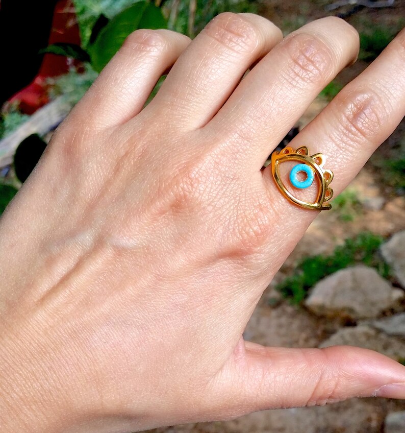 May include: Gold ring with a blue evil eye design. The ring is worn on a person's finger.
