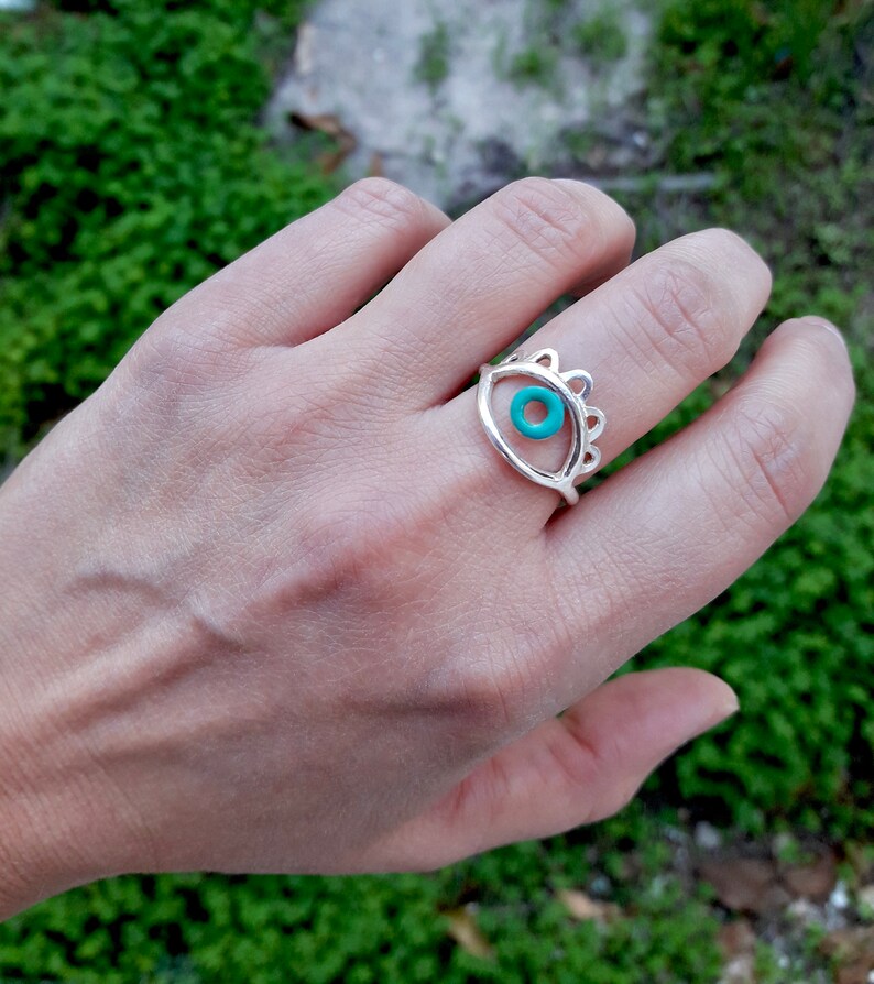 May include: Silver ring with a turquoise eye design. The ring is on a person's finger.