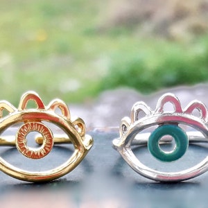 May include: Two eye rings, one gold and one silver, with a detailed eye design. The gold ring has a plain eye, while the silver ring has a green eye.