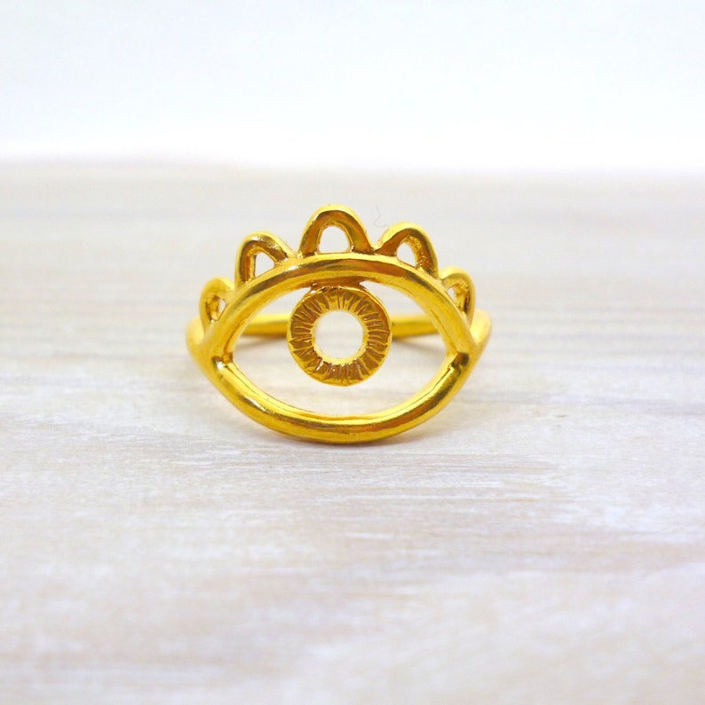 May include: Gold evil eye ring with a detailed design. The ring is made of a single piece of metal and has a raised eye shape with a smaller circle in the center.