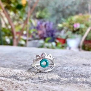 May include: Silver ring with a turquoise evil eye charm. The ring is designed to look like an eye with a crown of small points above it.