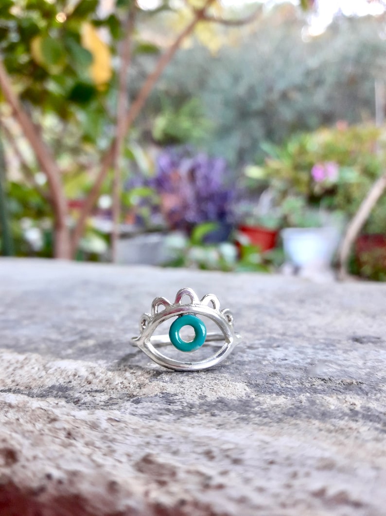 May include: Silver ring with a turquoise evil eye charm. The ring is designed to look like an eye with a crown of small points above it.