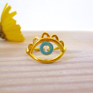 May include: Gold ring with a blue evil eye design. The ring is on a light wood surface.