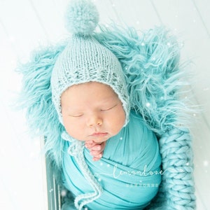 May include: A newborn baby wrapped in a turquoise blanket and wearing a light blue knitted hat with a pom-pom. The baby is nestled in a fluffy, turquoise blanket inside a wooden box. The image has a soft, dreamy aesthetic.