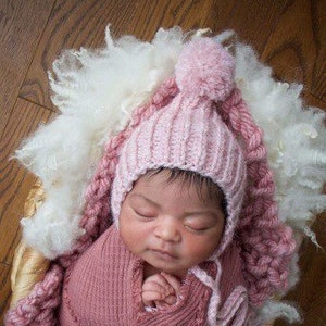 Könnte beinhalten: Ein neugeborenes Baby, eingewickelt in eine rosa Strickdecke und mit einer passenden rosa Mütze mit Bommel. Das Baby liegt in einem geflochtenen Korb mit weißem, flauschigem Material. Der Hintergrund ist eine Holzoberfläche.
