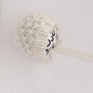May include: A cream-colored knitted baby bonnet with a textured pattern. The bonnet is tied with long, thin strings. The bonnet covers a white and black patterned object.