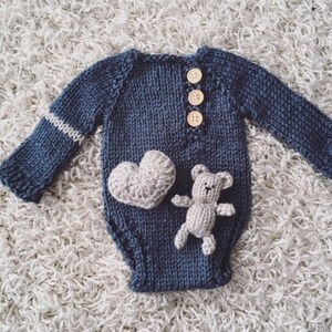 May include: A navy blue knitted baby onesie with long sleeves. The onesie features three wooden buttons, a white stripe on the left sleeve, and a gray crocheted heart and teddy bear applique. The garment is designed for newborns.