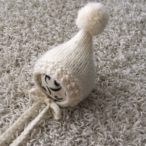 Könnte beinhalten: Eine weiße gestrickte Babyhaube mit einem Bommel oben und Bändern. Die Haube ist aus weichem, flauschigem Garn.