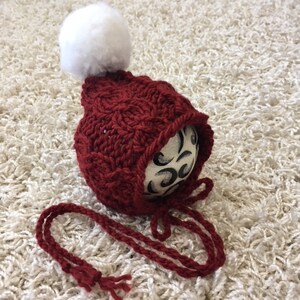 Peut inclure: Un bonnet de bébé en crochet rouge avec un pompon blanc et des liens assortis. Le bonnet est en point de câble.