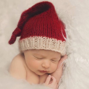 May include: A sleeping newborn wearing a red and cream knitted hat with a long, pointed end, resting on a fluffy white surface. The hat is a festive colour, and the baby's face is serene.
