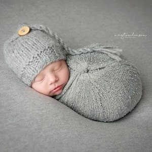Op de afbeelding: Een pasgeboren baby is gewikkeld in een grijze gebreide doek en draagt een grijze gebreide muts met een houten knoop. De baby slaapt rustig.