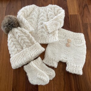 May include: A white knitted baby outfit set including a sweater, hat, and mittens. The hat has a fluffy pompom on top.