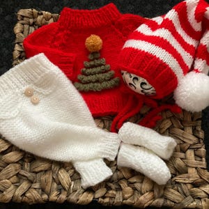 May include: A festive Christmas-themed baby outfit set. The set includes a red knitted sweater with a green Christmas tree applique, a white knitted bottom with two wooden buttons, a red and white striped hat with a white pom-pom, and white knitted mittens.
