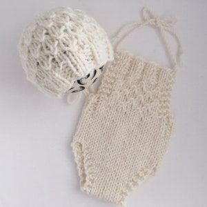 May include: A white knitted baby outfit with a matching hat. The outfit is a sleeveless romper with a scalloped edge. The hat is a simple beanie style.