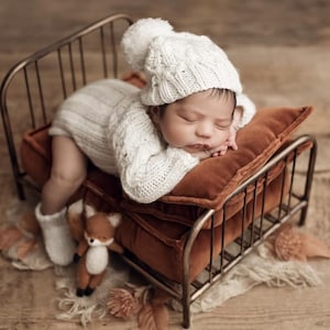 Op de afbeelding: Een pasgeboren baby met een witte gebreide muts met een pompon en bijpassende trui, liggend op een klein bed in antieke stijl met een roestkleurig kussen. Een kleine knuffelvos en sokken liggen aan de voeten van de baby. Het bed staat op een houten ondergrond.