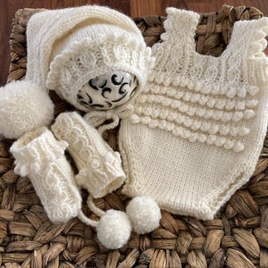 May include: A cream-colored, hand-knitted baby set, including a bonnet, mittens, romper, and two pom-poms. The set is made of soft yarn and features textured patterns. The romper has a round neckline and a series of bobbles.