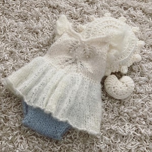May include: A white knitted baby outfit with a ruffled skirt and a matching hat. The outfit is made of a soft, delicate yarn and has a delicate lace pattern.