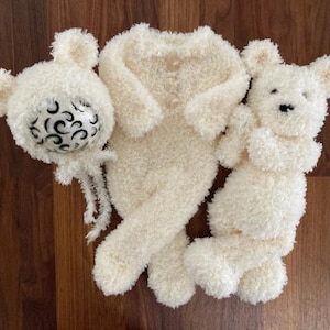 May include: A set of three white crocheted teddy bears. One bear is wearing a hat with a black and white swirl design. The bears are lying on a wooden surface.