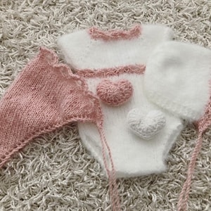 May include: A white knitted baby outfit with pink trim and two heart-shaped embellishments. The outfit includes a matching hat and a separate pink knitted bonnet.